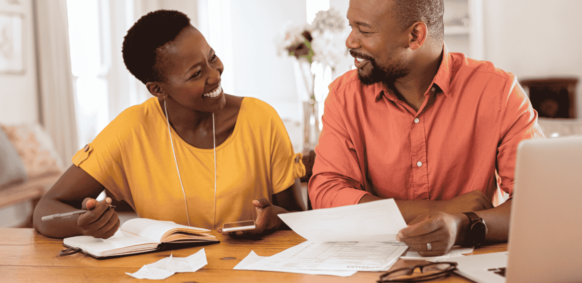 A smiling couple sits at a table with papers, a notebook, a pen, and a laptop, appearing to discuss finances or plan their travel budget together in a cozy home setting.