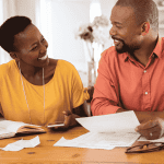 A smiling couple sits at a table with papers, a notebook, a pen, and a laptop, appearing to discuss finances or plan their travel budget together in a cozy home setting.
