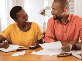 A smiling couple sits at a table with papers, a notebook, a pen, and a laptop, appearing to discuss finances or plan their travel budget together in a cozy home setting.