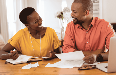 A smiling couple sits at a table with papers, a notebook, a pen, and a laptop, appearing to discuss finances or plan their travel budget together in a cozy home setting.