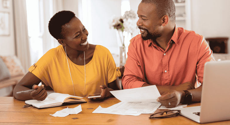 A smiling couple sits at a table with papers, a notebook, a pen, and a laptop, appearing to discuss finances or plan their travel budget together in a cozy home setting.
