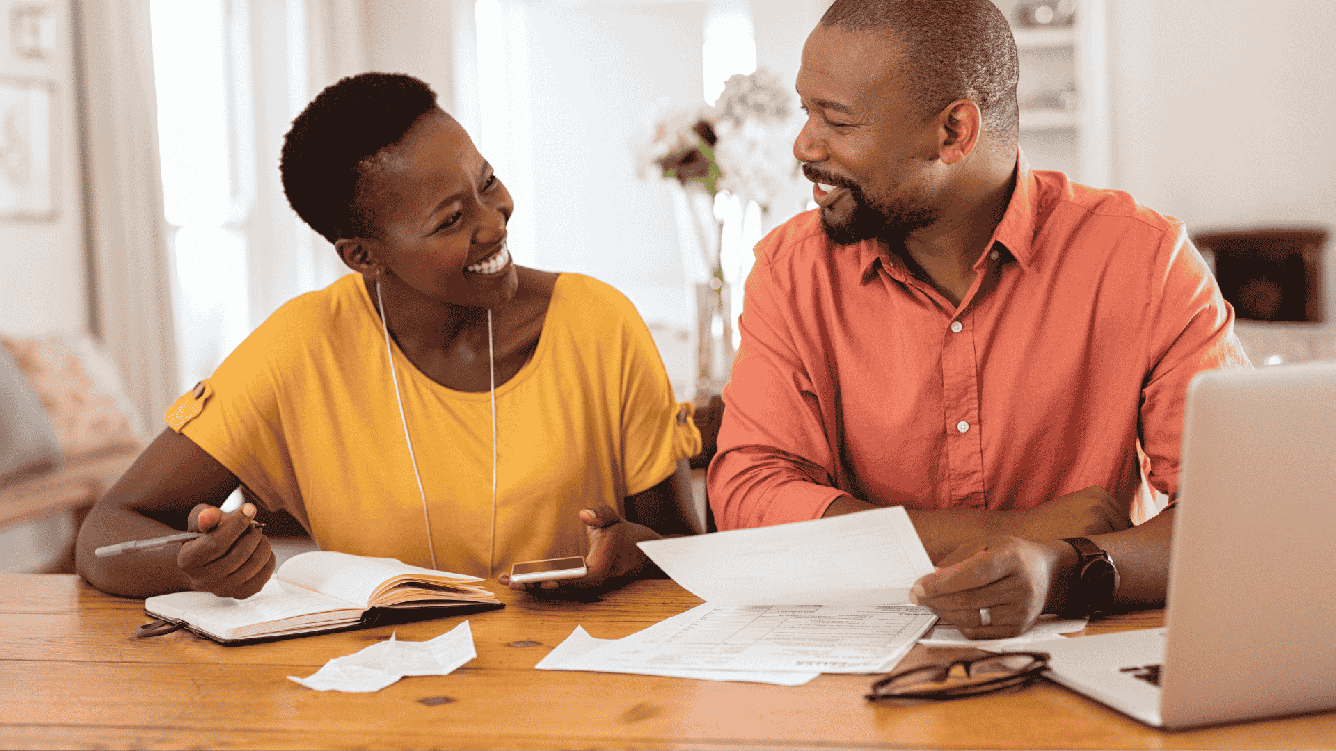 A smiling couple sits at a table with papers, a notebook, a pen, and a laptop, appearing to discuss finances or plan their travel budget together in a cozy home setting.