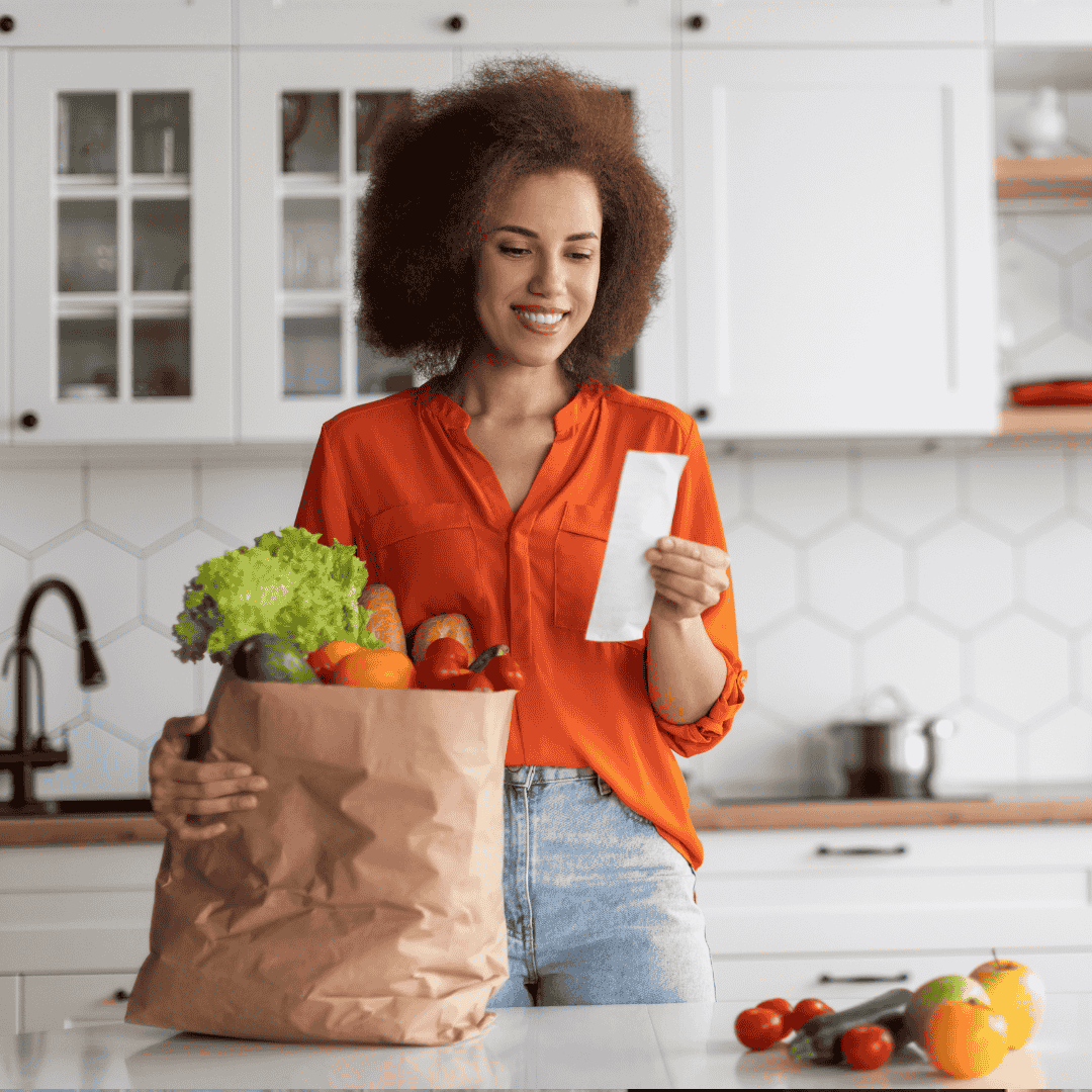 A smiling woman in an orange shirt stands in a kitchen, holding a grocery receipt in one hand and a paper bag full of fresh vegetables in the other, looking ready to cook up flavors inspired by her recent travel adventures.