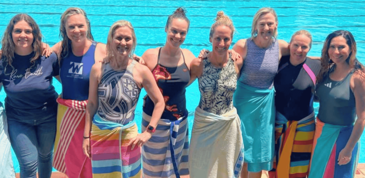 Eight women stand side by side in front of a swimming pool, smiling at the camera. Most are wearing swimsuits and have colorful towels wrapped around their waists, enjoying some fuel relief on a sunny day.