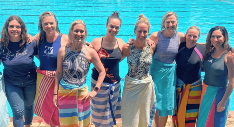 Eight women stand side by side in front of a swimming pool, smiling at the camera. Most are wearing swimsuits and have colorful towels wrapped around their waists, enjoying some fuel relief on a sunny day.