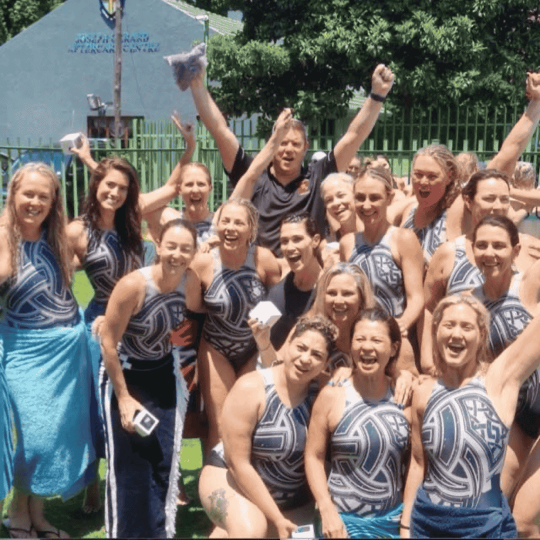 A group of smiling women in matching patterned swimsuits pose together outdoors, some holding white cards celebrating fuel relief. Most are cheering with raised arms, and green trees and a building are visible in the background.