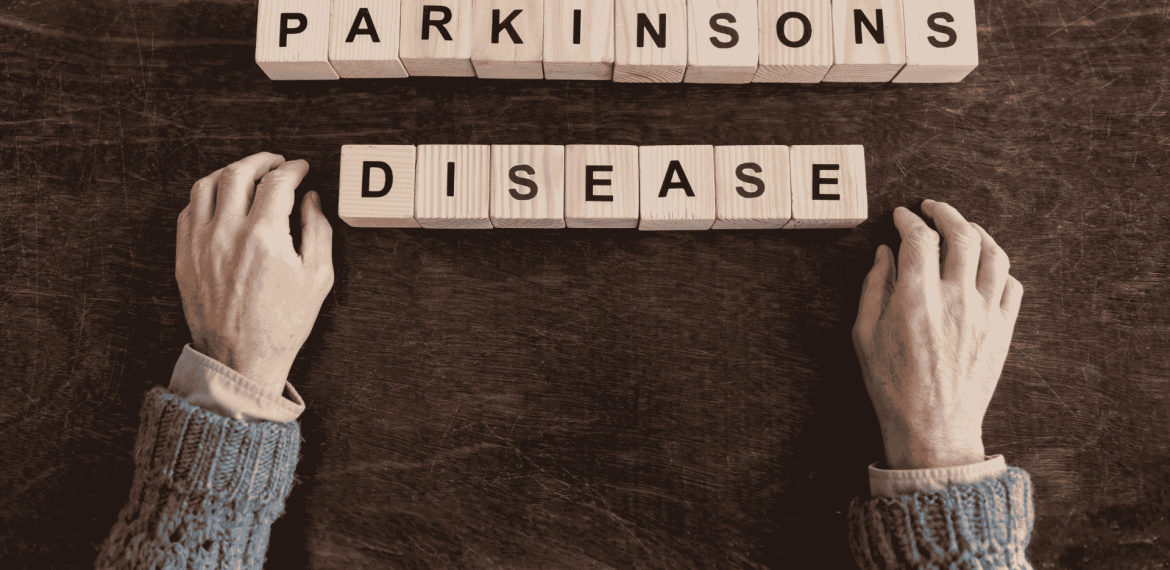Two hands rest on a dark surface near wooden blocks spelling out PARKINSONS DISEASE, suggesting the search for fuel relief and hope. The hands are positioned below the words in this sepia-toned image.