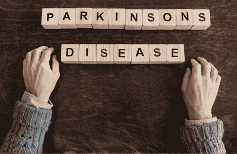 Two hands rest on a dark surface near wooden blocks spelling out PARKINSONS DISEASE, suggesting the search for fuel relief and hope. The hands are positioned below the words in this sepia-toned image.