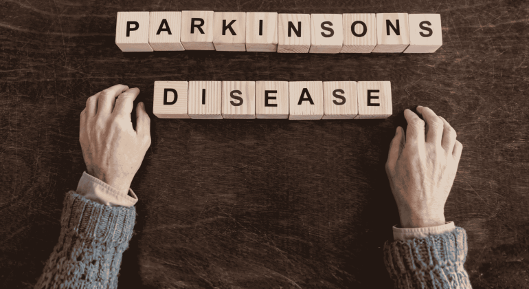 Two hands rest on a dark surface near wooden blocks spelling out PARKINSONS DISEASE, suggesting the search for fuel relief and hope. The hands are positioned below the words in this sepia-toned image.