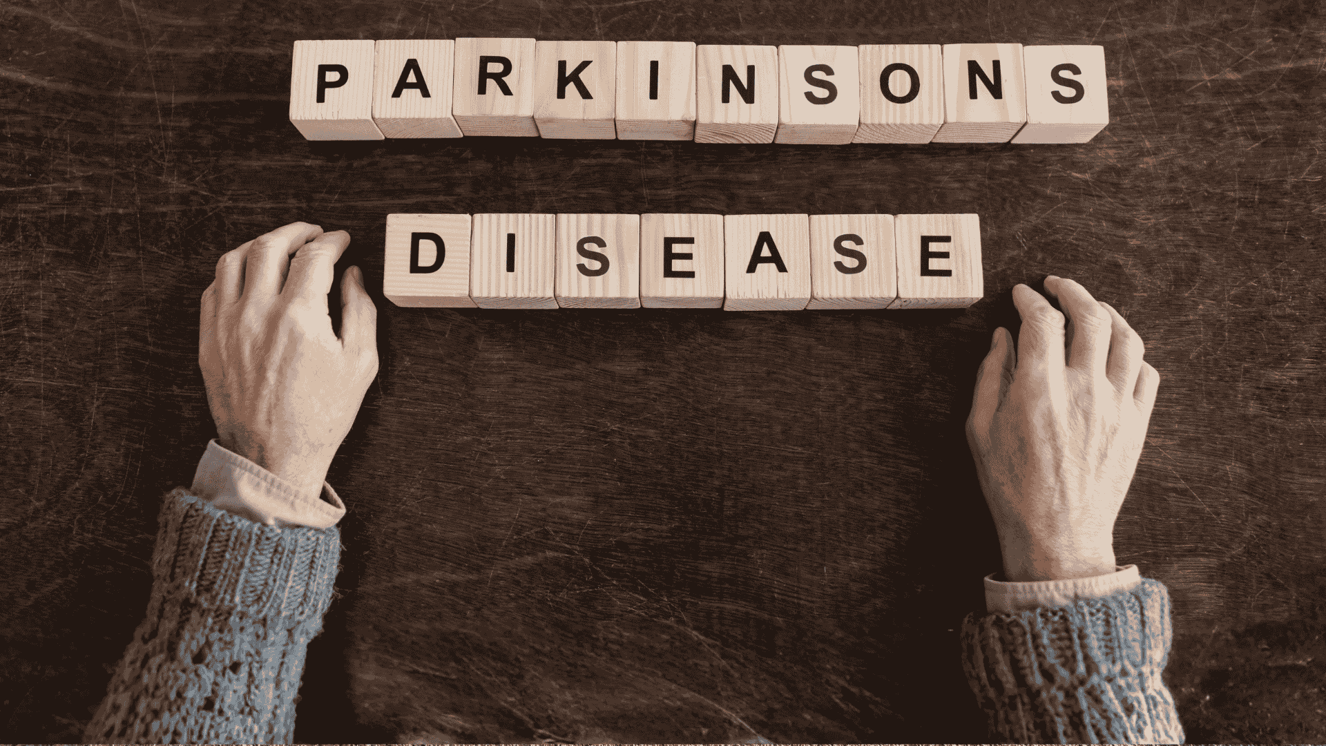 Two hands rest on a dark surface near wooden blocks spelling out PARKINSONS DISEASE, suggesting the search for fuel relief and hope. The hands are positioned below the words in this sepia-toned image.