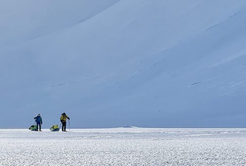 Two people in winter gear pull sleds across the vast, snowy landscape near Middelpos, with snow-covered slopes and a clear, open sky as a breathtaking backdrop.