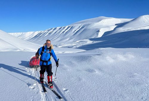 A person in winter gear is skiing across a snowy landscape near Middelpos, pulling a red sled under a clear blue sky with snow-covered mountains in the background.
