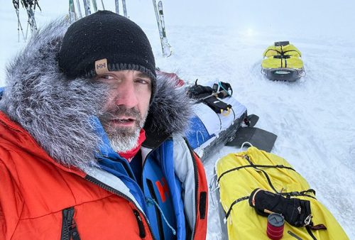 A man in winter gear with a fur-lined hood takes a selfie in the snowy Middelpos landscape. Behind him are sleds loaded with gear and several pairs of skis stuck upright in the snow.