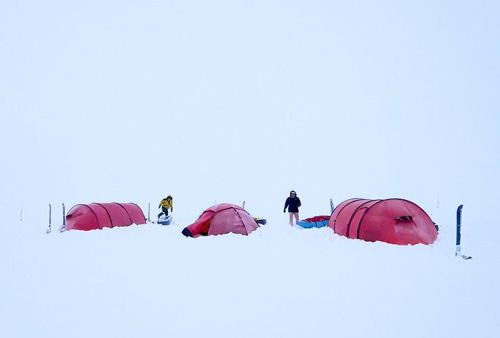 Three red tents are set up in a snowy, white Middelpos landscape. Two people stand near the tents, dressed in winter gear, surrounded by deep snow under an overcast sky.