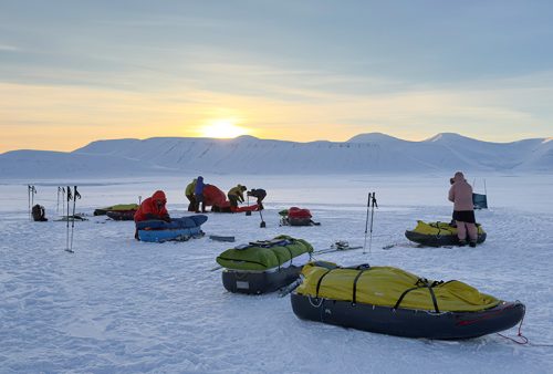 People in winter gear and one person in shorts stand on a snowy Middelpos landscape with sleds and equipment under a setting sun, snow-covered mountains rising in the background.