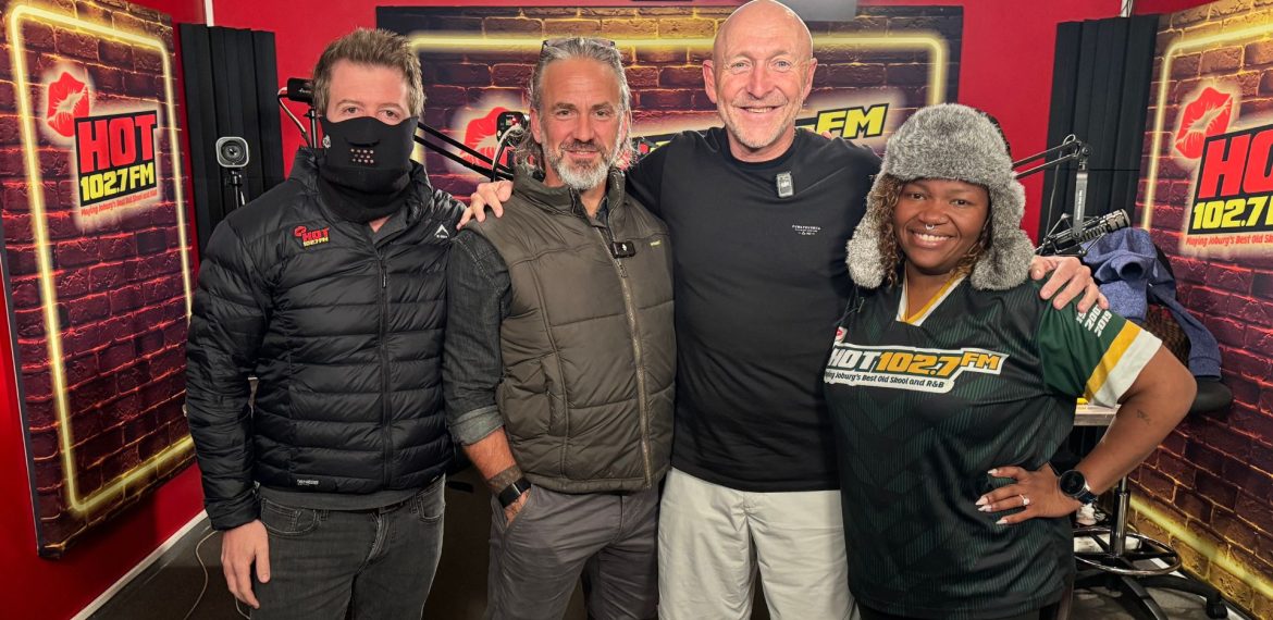 Four people stand together smiling in a HOT 102.7 FM radio studio, chatting about Middelpos. One wears a black mask and jacket, two men sport casual outdoor attire, and a woman dons a furry hat and station-branded shirt against red walls and microphones.