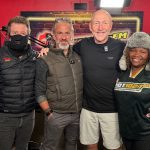 Four people stand together smiling in a HOT 102.7 FM radio studio, chatting about Middelpos. One wears a black mask and jacket, two men sport casual outdoor attire, and a woman dons a furry hat and station-branded shirt against red walls and microphones.