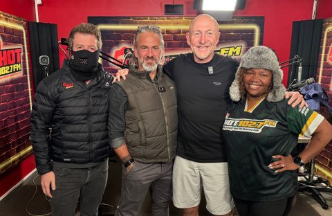 Four people stand together smiling in a HOT 102.7 FM radio studio, chatting about Middelpos. One wears a black mask and jacket, two men sport casual outdoor attire, and a woman dons a furry hat and station-branded shirt against red walls and microphones.