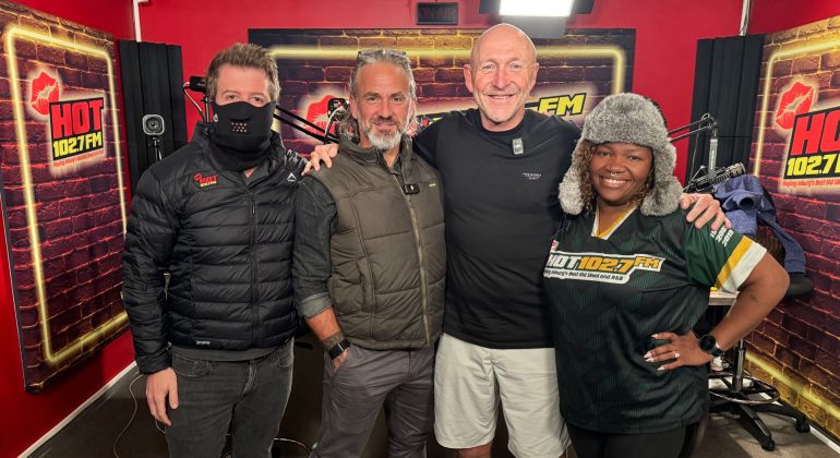 Four people stand together smiling in a HOT 102.7 FM radio studio, chatting about Middelpos. One wears a black mask and jacket, two men sport casual outdoor attire, and a woman dons a furry hat and station-branded shirt against red walls and microphones.