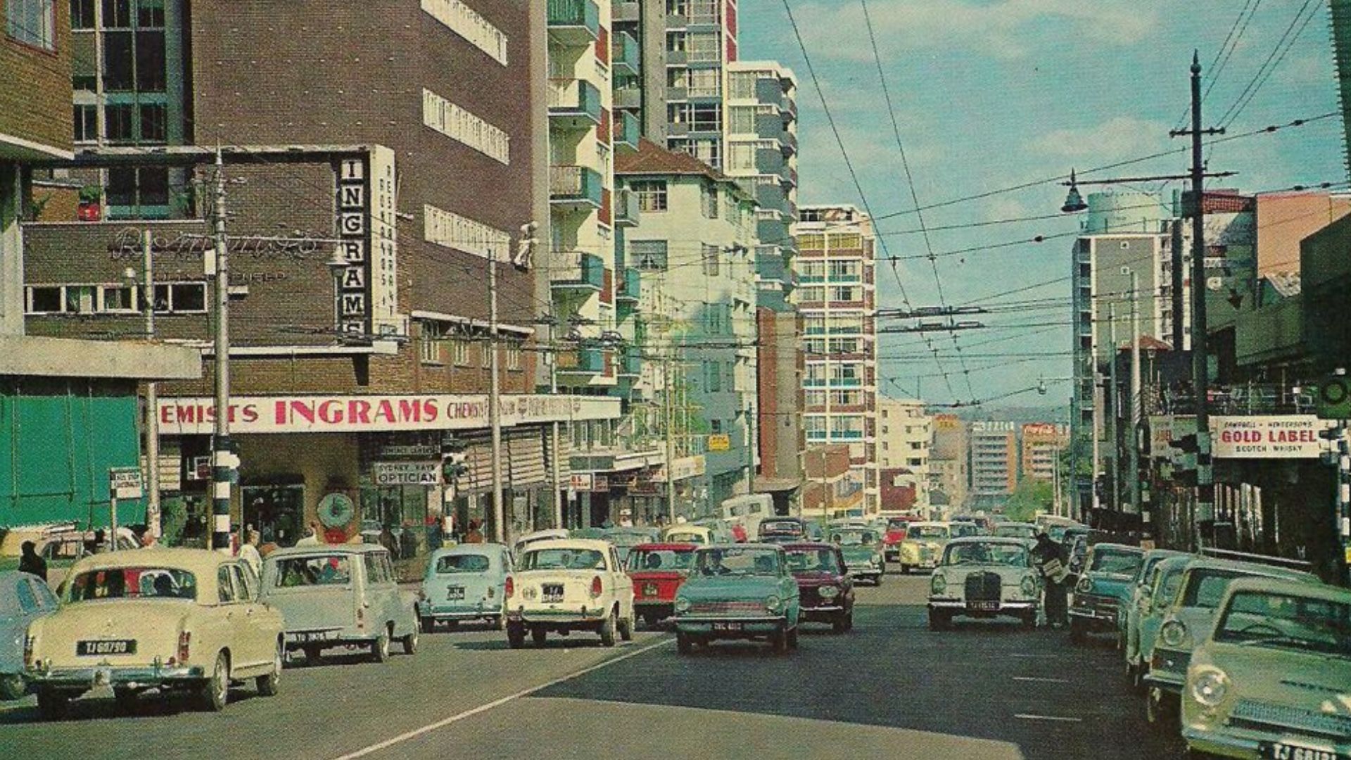 A busy city street scene from the mid-20th century with vintage cars, people walking on sidewalks, tall buildings, and shop signs—including Ingrams and Pet TV—under a clear blue sky.