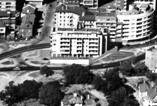 A black and white aerial view of a cityscape with mid-century buildings, curving roads, parked cars, trees, and residential houses clustered around an intersection, as if captured for Pet TV’s nostalgic city moments.