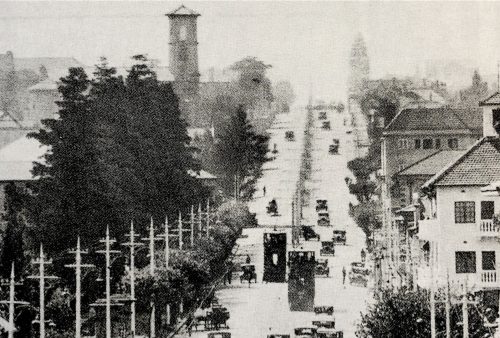Black and white photo of a busy city street lined with utility poles and trees, early 20th-century cars and rooftops; in the background, a tall clock tower stands above it all—capturing a scene reminiscent of classic films on Pet TV.