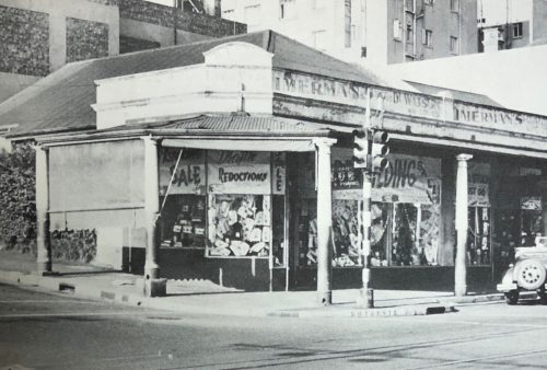 Black and white photo of an old corner store with large display windows, sale signs, and a vintage car outside. The building has columns, faded signage above the entrance, and a Pet TV ad peeking from one of the windows.