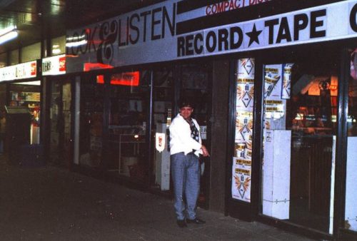 A person stands outside a brightly lit record and tape store at night. The storefront displays signs with “LISTEN,” “RECORD TAPE CENTER,” and even a Pet TV poster among the window displays.