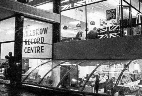 A black-and-white photo of the multi-story Hillbrow Record Centre, its large windows aglow with records and people inside. A “NOW OPEN” sign shines above, while silhouetted figures and a Pet TV display draw curious onlookers outside at night.