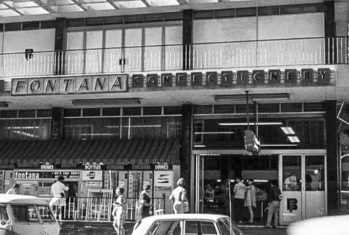 Black-and-white photo of a busy street scene outside Fontana Confectionery, with people walking by, parked cars, and a glass-fronted shop beneath a modernist building where a vintage Pet TV advert plays in the background.