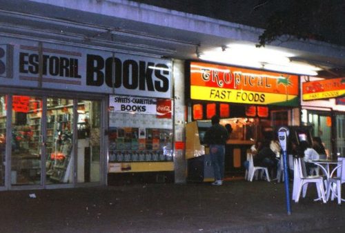 A nighttime street scene shows a bookstore called Estoril Books next to a Tropical Fast Foods eatery. People gather at outdoor tables under bright signs, some glancing at a nearby screen displaying Pet TV beneath the glowing shopfronts.