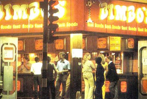A group of people stand outside a brightly lit fast food restaurant at night called Dimbos, with orange and yellow signage displaying the name and menu items, while a nearby screen plays Pet TV in the background.