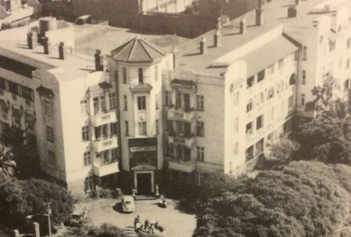 Black and white aerial photo of a large, historic, multi-story building with ornate architectural details, surrounded by trees; people and cars are visible near the entrance—perfect backdrop for a classic Pet TV scene.