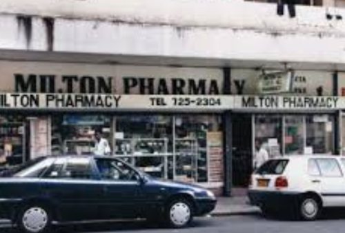 Two cars are parked in front of Milton Pharmacy, which features large windows displaying products—including a Pet TV in the corner—and a sign above the entrance with the business name and phone number.