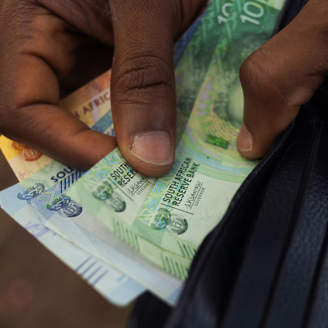 A close-up of a hand pulling several South African rand banknotes from a black wallet, with a Standard Bank card partially visible inside.