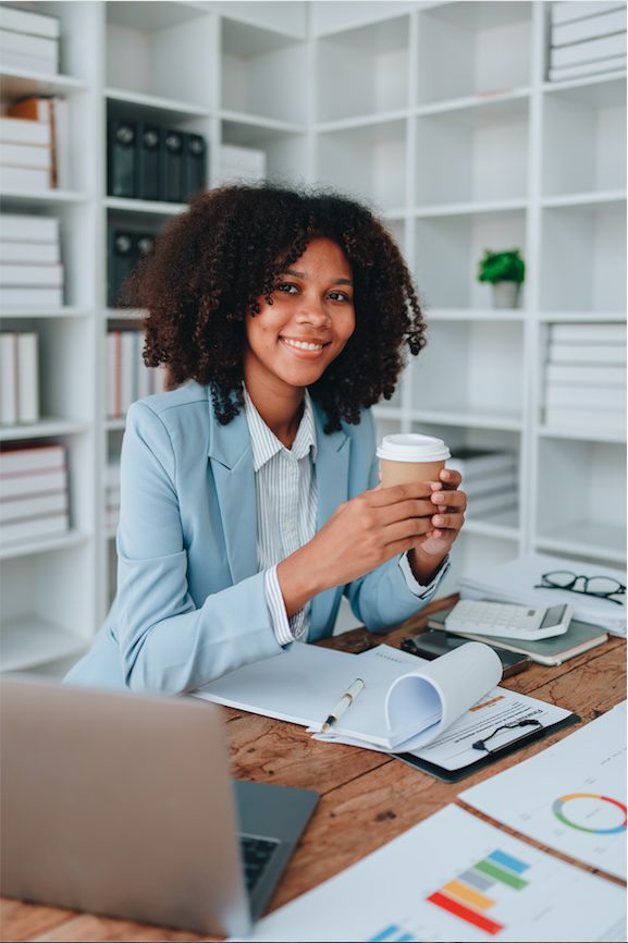 A woman in a light blue blazer sits at a desk with a laptop, documents, and charts, smiling and holding a coffee cup as she plans her next business travel in an office with shelves and binders in the background.