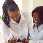 Two women in business attire smile as one sits and writes on a document while the other leans over, offering guidance—perhaps planning their next business travel—in a bright office with a laptop and a plant in the background.