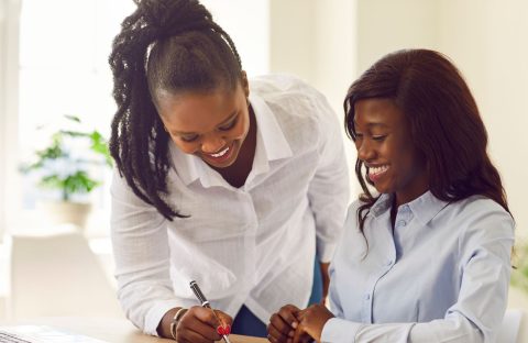 Two women in business attire smile as one sits and writes on a document while the other leans over, offering guidance—perhaps planning their next business travel—in a bright office with a laptop and a plant in the background.