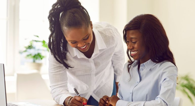 Two women in business attire smile as one sits and writes on a document while the other leans over, offering guidance—perhaps planning their next business travel—in a bright office with a laptop and a plant in the background.