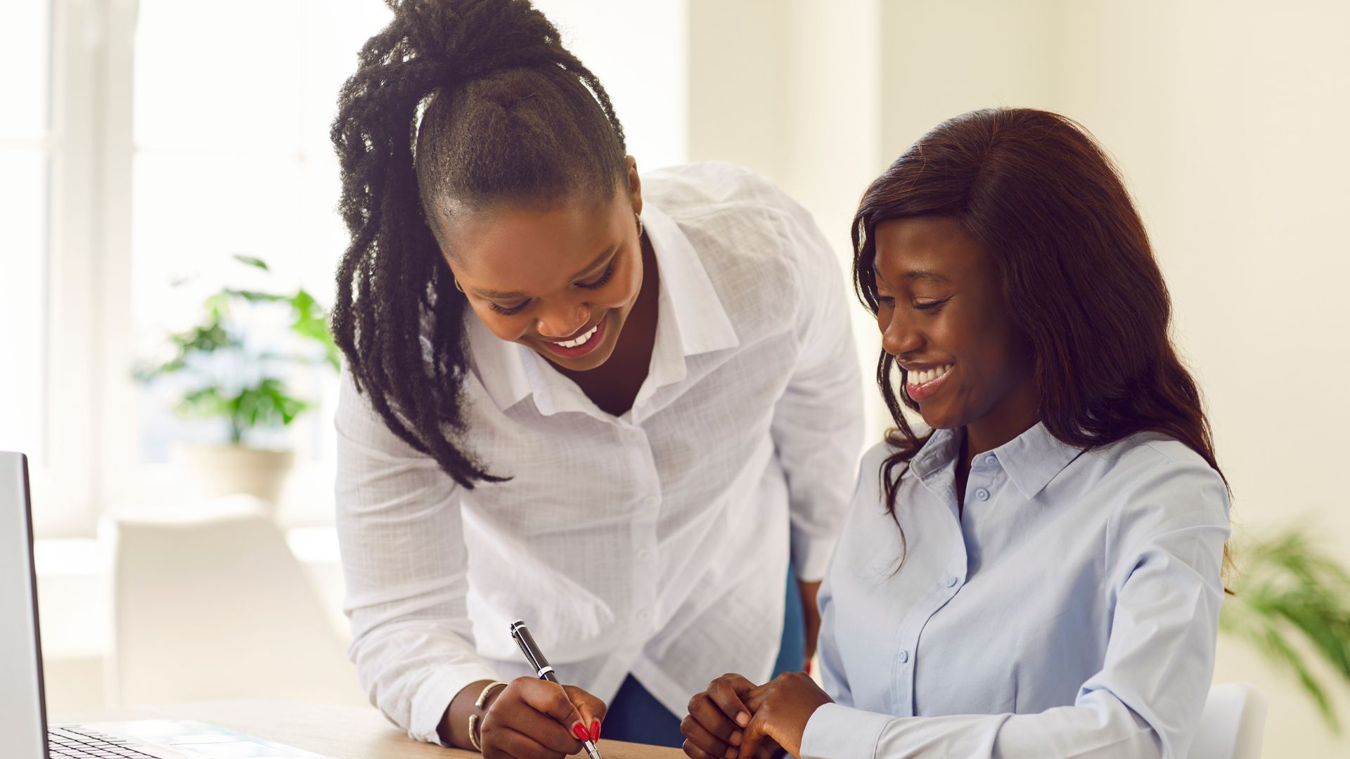 Two women in business attire smile as one sits and writes on a document while the other leans over, offering guidance—perhaps planning their next business travel—in a bright office with a laptop and a plant in the background.