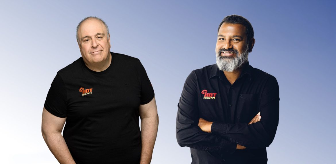 Two men stand before a gradient blue background, both wearing HOT Realty shirts. Lloyd Madurai, on the right in a black button-up shirt with arms crossed, smiles beside another man in a black t-shirt.