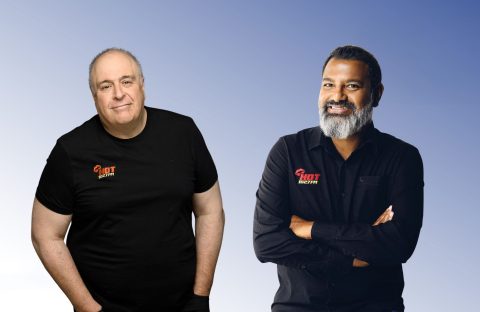 Two men stand before a gradient blue background, both wearing HOT Realty shirts. Lloyd Madurai, on the right in a black button-up shirt with arms crossed, smiles beside another man in a black t-shirt.