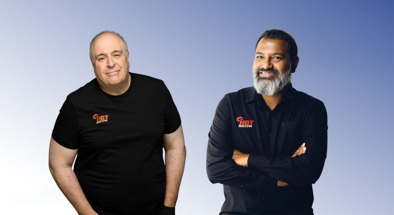 Two men stand before a gradient blue background, both wearing HOT Realty shirts. Lloyd Madurai, on the right in a black button-up shirt with arms crossed, smiles beside another man in a black t-shirt.