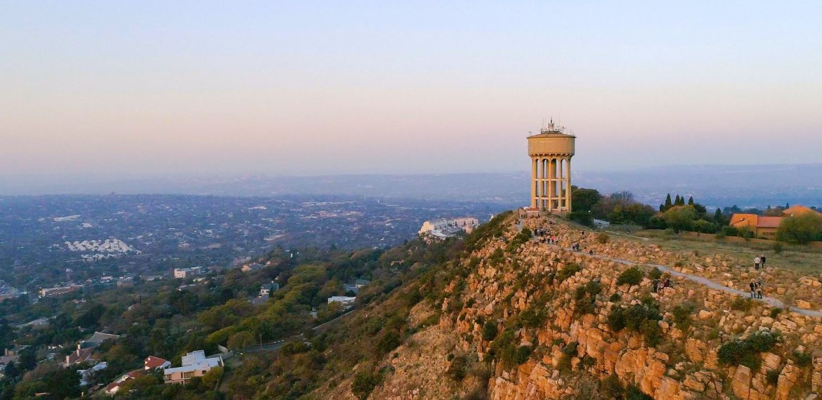 A tall water tower stands on a rocky hilltop overlooking a sprawling cityscape at sunset, with scattered houses and greenery below—a perfect scene for a Pet TV backdrop against the hazy evening sky.