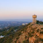 A tall water tower stands on a rocky hilltop overlooking a sprawling cityscape at sunset, with scattered houses and greenery below—a perfect scene for a Pet TV backdrop against the hazy evening sky.