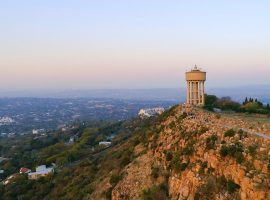 A tall water tower stands on a rocky hilltop overlooking a sprawling cityscape at sunset, with scattered houses and greenery below—a perfect scene for a Pet TV backdrop against the hazy evening sky.