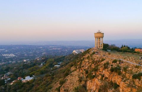 A tall water tower stands on a rocky hilltop overlooking a sprawling cityscape at sunset, with scattered houses and greenery below—a perfect scene for a Pet TV backdrop against the hazy evening sky.