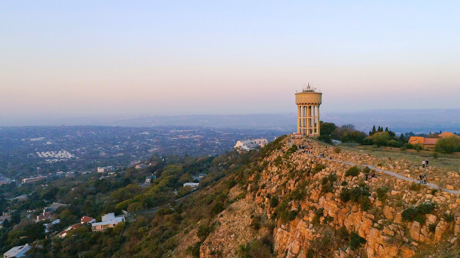 A tall water tower stands on a rocky hilltop overlooking a sprawling cityscape at sunset, with scattered houses and greenery below—a perfect scene for a Pet TV backdrop against the hazy evening sky.