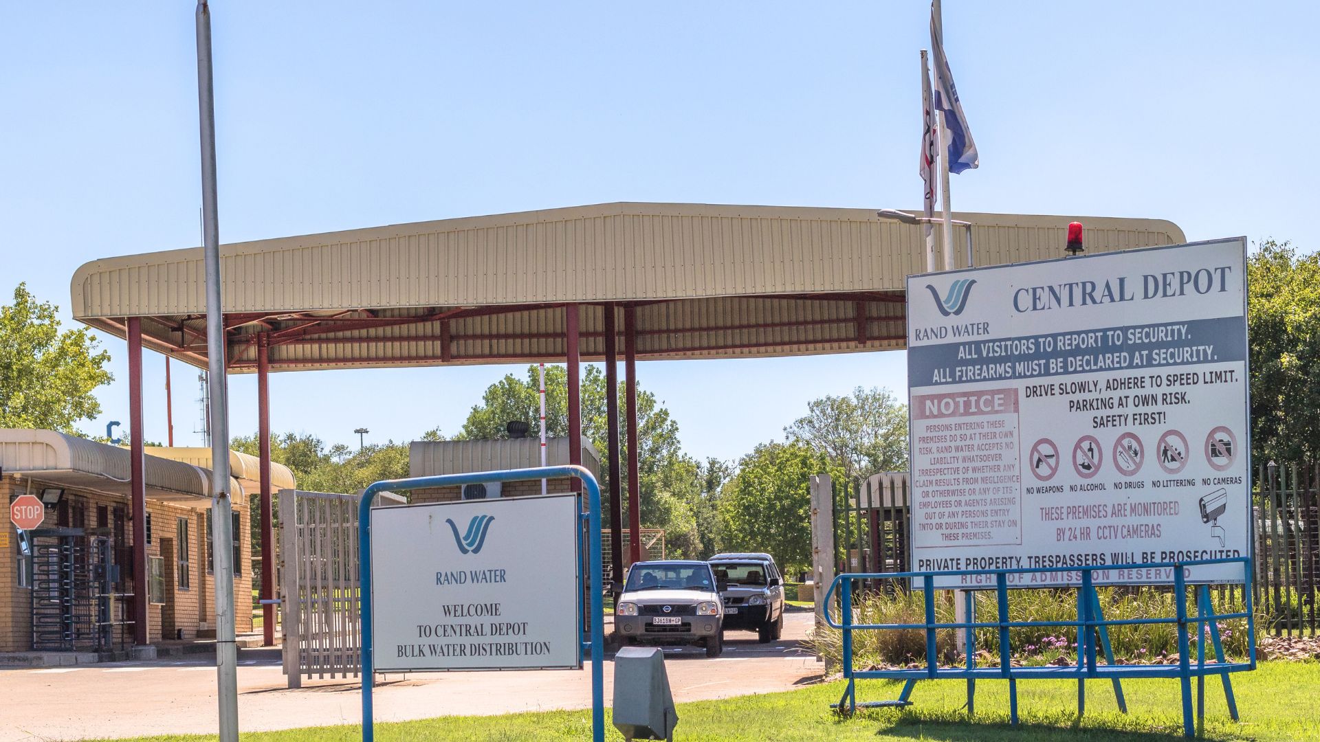 Entrance to Rand Water Central Depot with security gate, parked vehicles, two large signs displaying entry rules and information, and a stop sign on the left. Trees and a clear sky form the backdrop near Johannesburg water facilities.