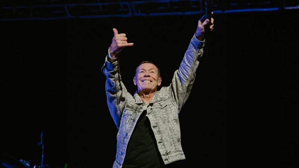 A man in a patterned jacket stands on stage with both arms raised, smiling and making a shaka hand sign, holding a microphone in one hand against a dark background, exuding confidence and the aura of wealth.